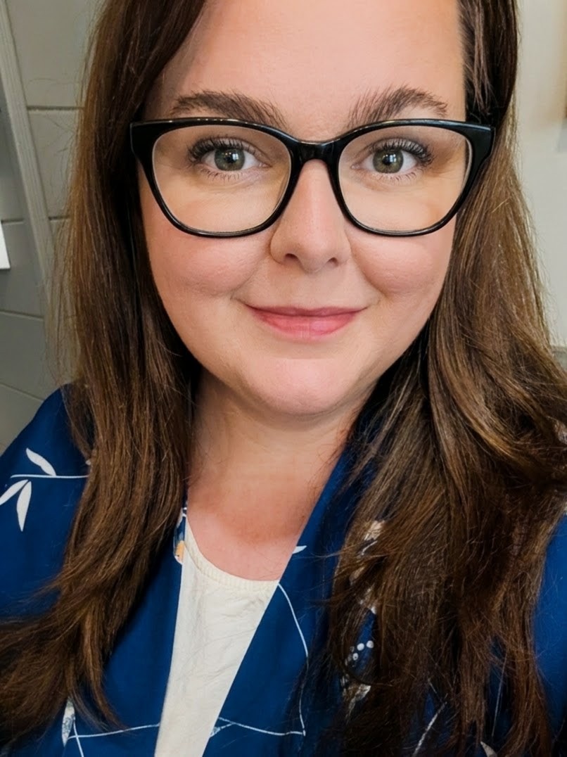 Jennifer Jackson with long brown hair, wearing black glasses, smiling, dressed in a blue patterned blazer over a cream-coloured top.